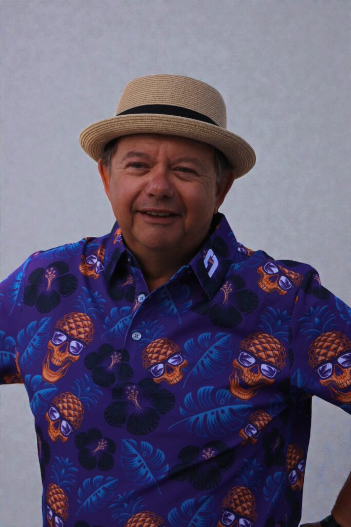 Un homme portant un chapeau de paille bronzé et une chemise d'un violet éclatant ornée de têtes de mort et d'ananas orange sourit sur un fond uni de couleur claire.
