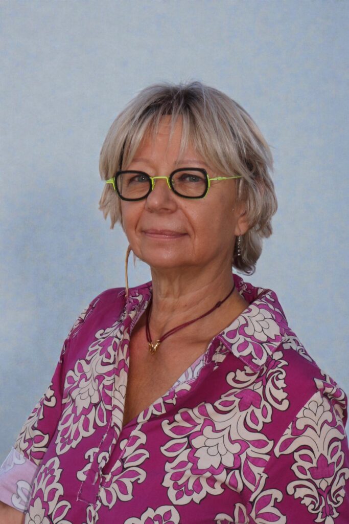Une femme aux cheveux blonds courts et aux lunettes à monture verte sourit légèrement. Elle porte une chemise rose et blanche à motifs floraux, un collier, et pose devant un fond bleu clair.