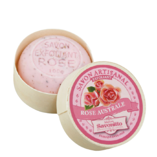 Un savon rond rose "Savon exfoliant parfum rose-100g" dans un récipient beige ouvert avec un motif floral de rose sur le couvercle, partiellement visible à l'intérieur de l'emballage.
