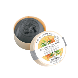 Savonitto Savon fleur d'oranger-100 g est un savon artisanal noir rond dans un contenant en bois clair, étiqueté "Savon Artisanal, Miel Fleur d'Oranger" et orné d'illustrations de fleurs et de nids d'abeilles.