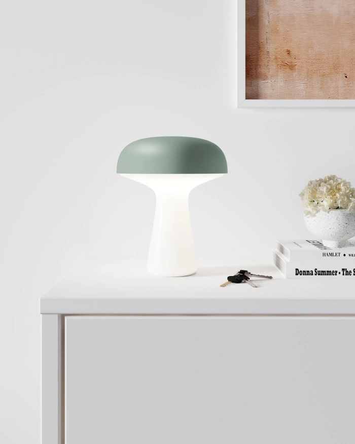 Une lampe de table moderne avec un abat-jour vert en forme de dôme et une base blanche est posée sur un meuble blanc, à côté d'une pile de livres, de fleurs blanches et d'un trousseau de clés ; une partie d'une œuvre d'art encadrée est visible au-dessus.