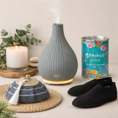 Un diffuseur d'arômes gris nervuré émet de la brume à côté d'une bougie allumée, d'une boîte de graines de fleurs, de chaussures noires, d'un coussin en tissu rayé avec une houppe, d'une natte ronde en paille et de branches d'eucalyptus vertes.