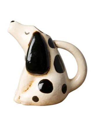 Le "Pichet chien grès 20,5 cm" est une théière en céramique en forme de chien assis avec des taches noires, de grandes oreilles noires, une poignée courbée en guise de queue et un bec verseur ressemblant au nez du chien, regardant vers le haut.