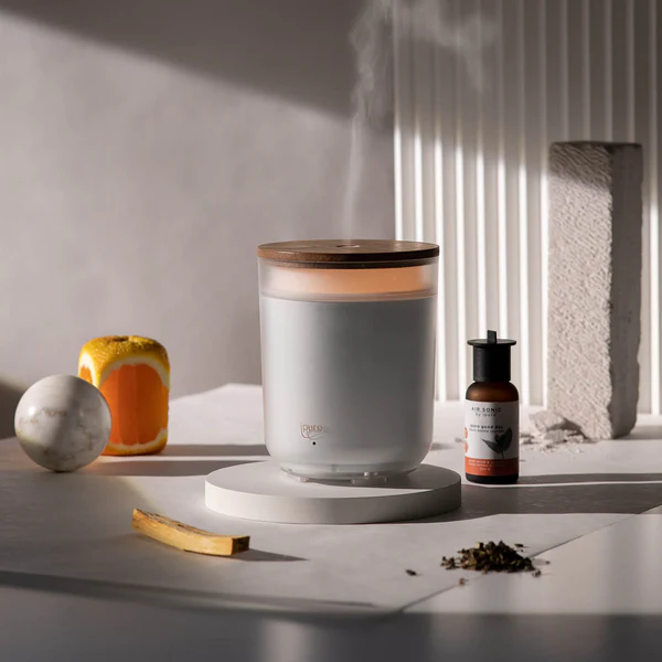 Le Diffuseur bougie ipuro, un diffuseur moderne d'huiles essentielles blanc avec un couvercle en bois sur une base ronde, émet de la vapeur au milieu d'un verre de boisson orange, d'un flacon compte-gouttes, d'une sphère en marbre, d'un bâton, de feuilles de thé et d'un bloc de béton texturé.