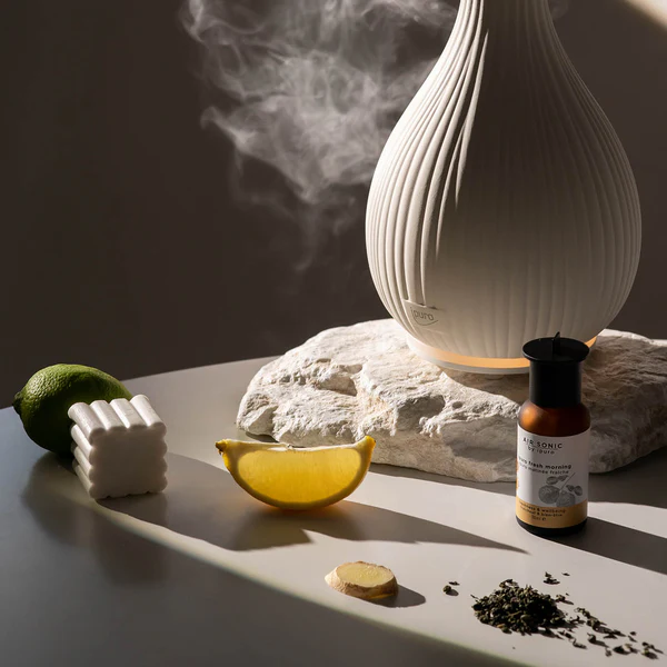 Le Diffuseur AIR SONIC Vase, un élégant diffuseur en céramique, émet de la brume à côté d'un quartier de citron, d'une tranche de gingembre, d'un citron vert, d'un petit bloc blanc, d'une bouteille brune et de feuilles de thé sur une pierre et une table dans une lumière douce et chaude.