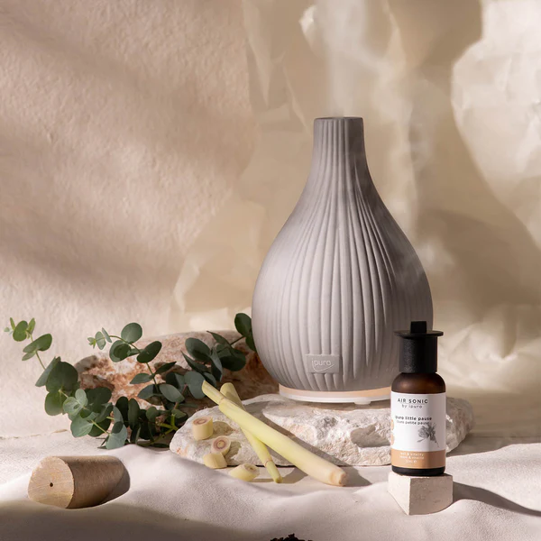 Le Diffuseur AIR SONIC Vase, un diffuseur d'aromathérapie en céramique grise, est présenté sur une pierre avec de l'huile essentielle, des tiges de citronnelle et des feuilles d'eucalyptus sur un fond doux et neutre.
