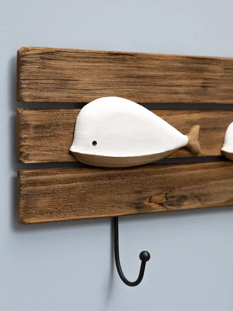 Le Porte-manteau baleines est un crochet mural en bois avec un décor de baleine blanc et beige sur des lattes rustiques, avec un crochet en métal noir en dessous, monté sur un mur bleu clair.