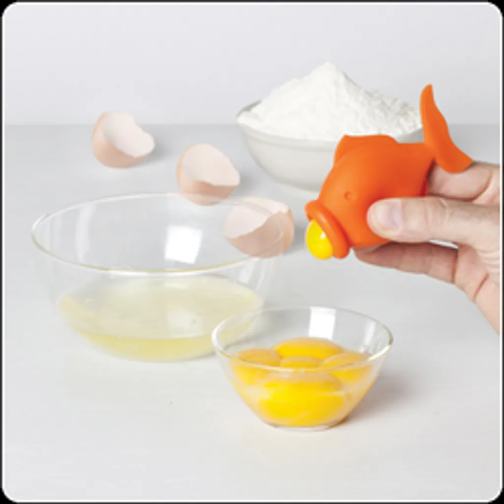 Une main utilise un outil orange en forme de poisson pour séparer les jaunes d'œufs des blancs d'œufs. Des coquilles d'œuf, un bol de farine, un bol avec des blancs d'œuf et un bol avec des jaunes d'œuf se trouvent sur une surface blanche.