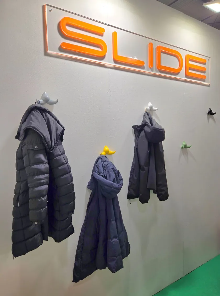 Quatre manteaux d'hiver sombres sont suspendus à des crochets à moustaches Patère Baffo, en forme de petites moustaches blanches, jaunes, vertes et noires, sous un panneau "SLIDE" orange vif sur un mur blanc.