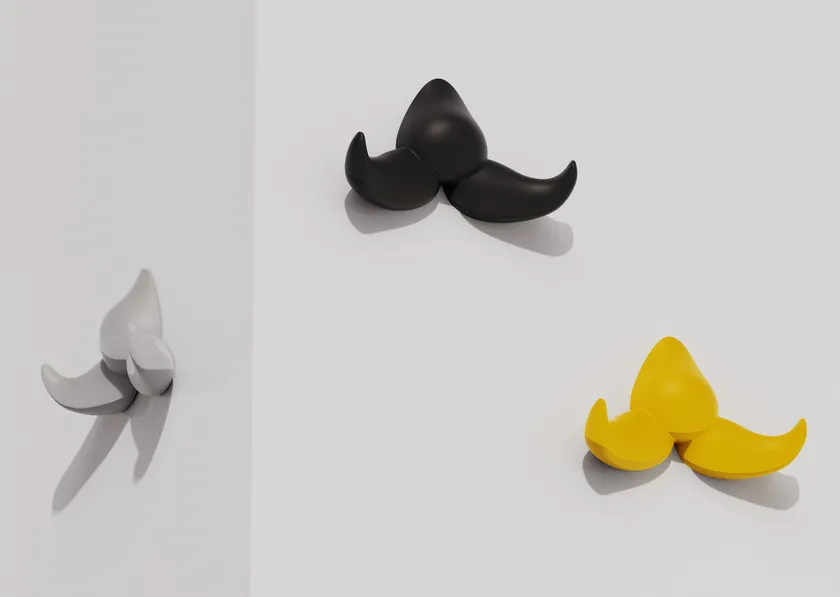 Trois crochets à moustache Patère Baffo gris, noir et jaune sont fixés sur un mur blanc, le crochet gris projetant une ombre sur la gauche.