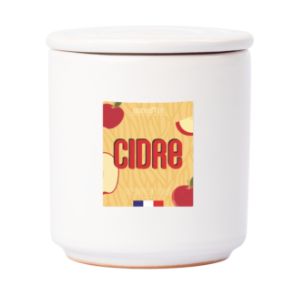 Bocal en céramique blanche avec couvercle, Bougie Cocktail Cidre présente une étiquette colorée indiquant "CIDRE" avec des pommes rouges, des tranches de pommes et un petit drapeau français au fond.
