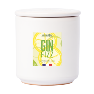 Un pot en céramique blanche avec couvercle, étiqueté "Bougie Cocktail Gin Fizz", comporte des illustrations de tranches de citron et de petits drapeaux français au fond.