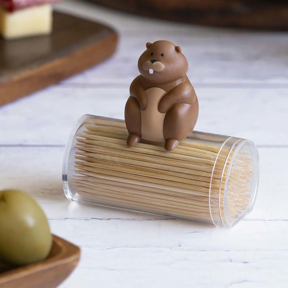 Le Distributeur cure-dents Woody est un récipient cylindrique transparent avec des cure-dents, surmonté d'une adorable figurine de castor de dessin animé de couleur marron. Il est posé sur une surface blanche, avec des aliments flous en arrière-plan.