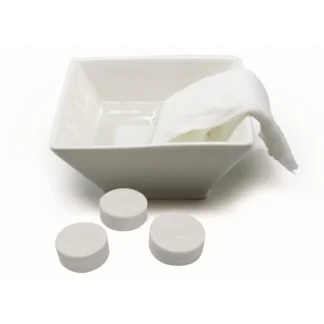 Un bol blanc carré rempli d'eau contient une serviette Lingettes Compressées Citron partiellement déroulée. Trois Lingettes Compressées Citron non utilisées sont placées devant le bol sur une surface blanche.