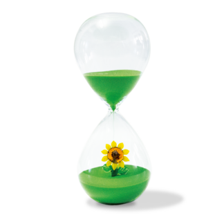 Le Sablier Verre 30 min présente du sable vert dans un sablier en verre. La partie supérieure contient le sable, tandis que la partie inférieure présente un tournesol jaune sur une base de sable vert. Fond blanc uni. Deux options de figurines sont disponibles.