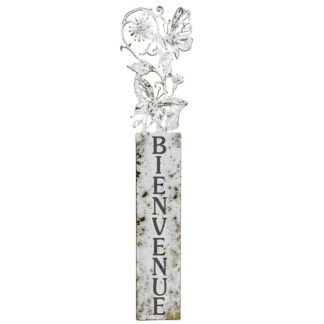 Une grande enseigne rustique en métal avec le mot "BIENVENUE" écrit verticalement en lettres noires et des fleurs décoratives et un papillon au sommet.