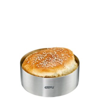 Un petit pain rond aux graines de sésame est placé à l'intérieur d'un moule à anneaux Moule Burger BBQ en acier inoxydable, portant le nom de la marque "GEFU" sur la face avant.