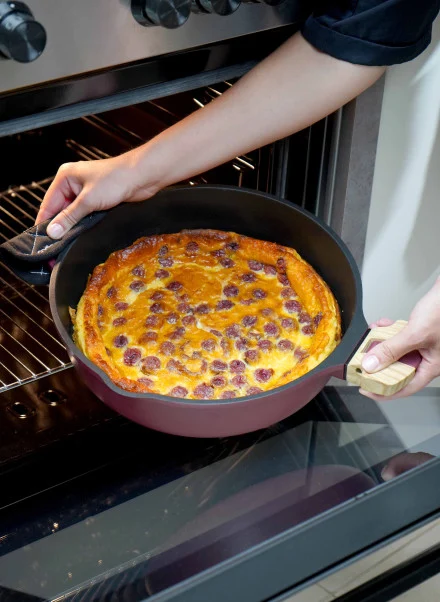 Avec des gants de cuisine, une personne sort du four un clafoutis aux cerises fraîchement cuit, à la croûte dorée et aux cerises apparentes, dans la Poêle Fabuleuse 28 cm.