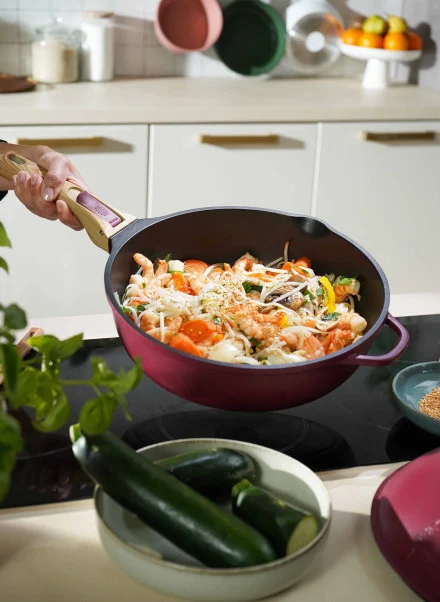 Quelqu'un prépare un sauté vibrant de crevettes, de légumes et de nouilles dans une poêle Poêle Fabuleuse 28 cm de couleur pourpre sur la cuisinière ; des courgettes fraîches et des herbes aromatiques se trouvent à proximité.