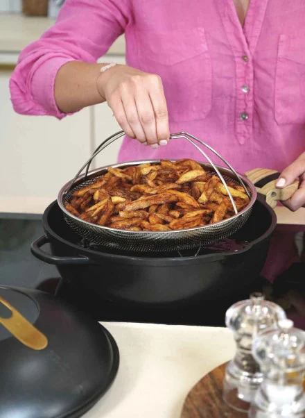 Une personne en chemise rose soulève un panier de quartiers de pommes de terre cuits de la Poêle Fabuleuse 28 cm sur une cuisinière, avec des salières et des poivrières à proximité.