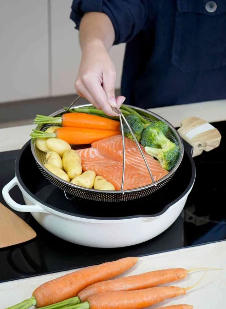 Avec la Poêle Fabuleuse 28 cm, une personne ajoute un panier à vapeur contenant du saumon, des pommes de terre grelots, des carottes et des brocolis dans la marmite blanche de la cuisinière pour faire cuire le poisson et les légumes.