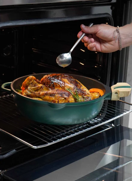 Une main utilise une cuillère pour arroser un poulet rôti avec des légumes dans une Poêle Fabuleuse verte de 28 cm, à l'intérieur d'un four ouvert.