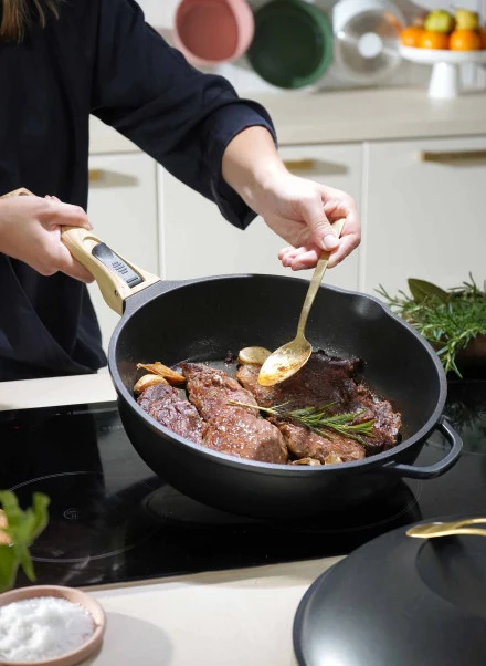Une personne fait cuire des steaks avec de l'ail et du romarin dans une poêle en fonte Poêle Fabuleuse de 28 cm sur la cuisinière, en arrosant la viande à l'aide d'une cuillère. Des bols et une corbeille de fruits sont posés sur le comptoir de la cuisine à l'arrière-plan.