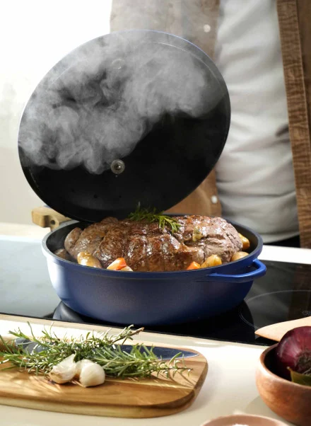 Un rôti fumant avec des légumes et des herbes se révèle à l'intérieur d'une Poêle Fabuleuse 28 cm sur la cuisinière, avec un brin de romarin frais à côté sur une planche à découper en bois.