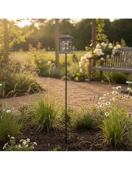 La Lanterne Jardin de Tuteur, une lanterne décorative sur un haut piquet métallique, illumine un jardin paisible rempli de fleurs sauvages et d'herbes, avec un banc en bois à l'arrière-plan créant une scène accueillante.