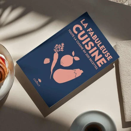 Le livre de cuisine "Livre Recettes Fabuleuse", dont la couverture est illustrée de carottes, de citrons et d'aubergines, est posé sur une table à côté d'une assiette et d'une serviette blanche, sous un soleil doux.