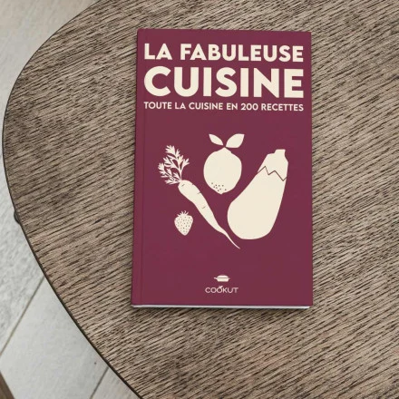 Sur une table ronde en bois trône un livre de cuisine intitulé "Livre Recettes Fabuleuse". Sa couverture est bordeaux, décorée d'illustrations beiges de légumes et de fruits.