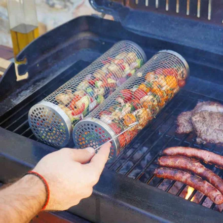 Le panier en filet RollGrill Barbecue Inox, rempli de brochettes de légumes et de viande, peut être tourné par une personne sur la grille d'un barbecue, à côté de saucisses et de steaks.