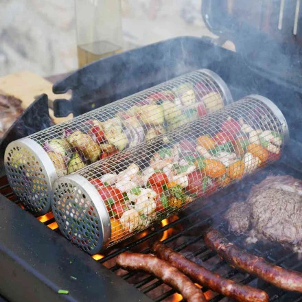 Deux paniers métalliques RollGrill Barbecue Inox remplis de légumes hachés et de crevettes sont posés sur un gril à côté de saucisses et de steaks, la fumée s'élevant des aliments grésillants.