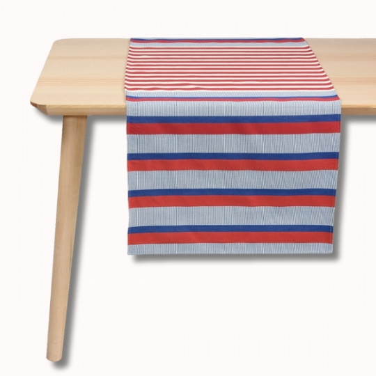 Une table en bois clair avec un chemin de table à rayures horizontales rouges, bleues et blanches. Le chemin de table comporte des rayures rouges et blanches sur le dessus et des rayures plus larges bleues, blanches et rouges qui pendent sur le bord de la table.