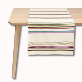 Une table en bois clair avec un chemin de table de couleur crème comportant des rayures horizontales violettes, jaunes, vertes, noires et rouges. Le chemin de table est soigneusement placé au centre de la table.