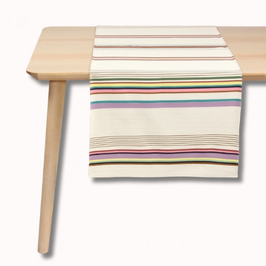 Une table en bois clair avec un chemin de table de couleur crème comportant des rayures horizontales violettes, jaunes, vertes, noires et rouges. Le chemin de table est soigneusement placé au centre de la table.