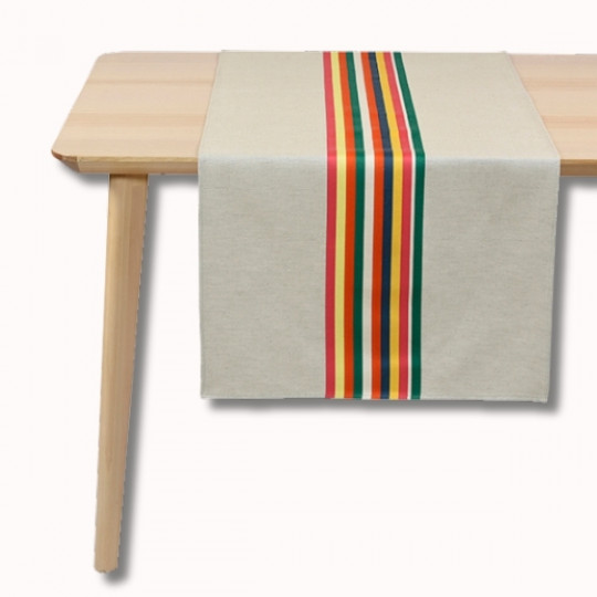Une table en bois clair avec un chemin de table de couleur crème avec des rayures verticales rouges, jaunes, vertes et orange au centre.