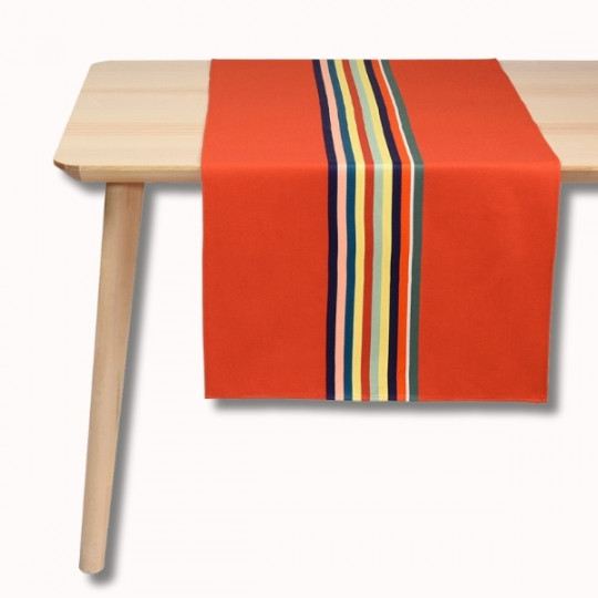Une table en bois clair avec un chemin de table orange à rayures verticales multicolores au centre. Le fond est blanc.