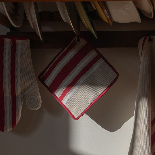 Un gant de cuisine et un cache-pot rayés rouge et blanc sont suspendus à des crochets sous une étagère en bois où sont empilées des assiettes. La lumière du soleil projette des ombres sur le mur.
