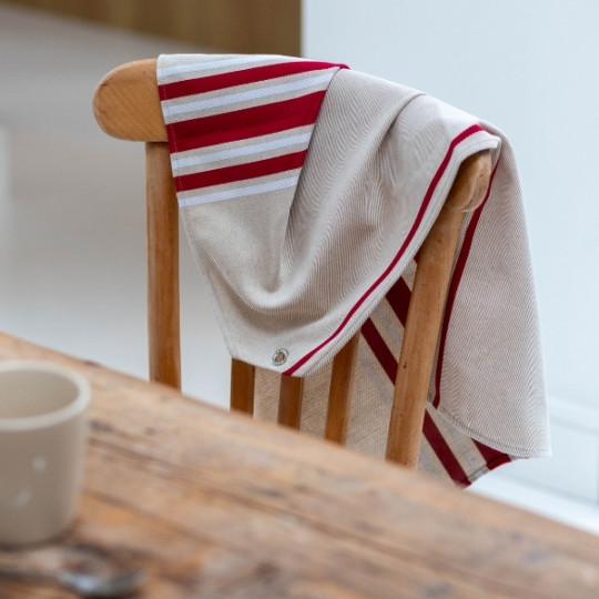 Le Torchon 70x53 (6 coloris), un tissu beige avec des rayures rouges et blanches, est drapé sur une chaise en bois à côté d'une table en bois, avec une tasse floue visible au premier plan.