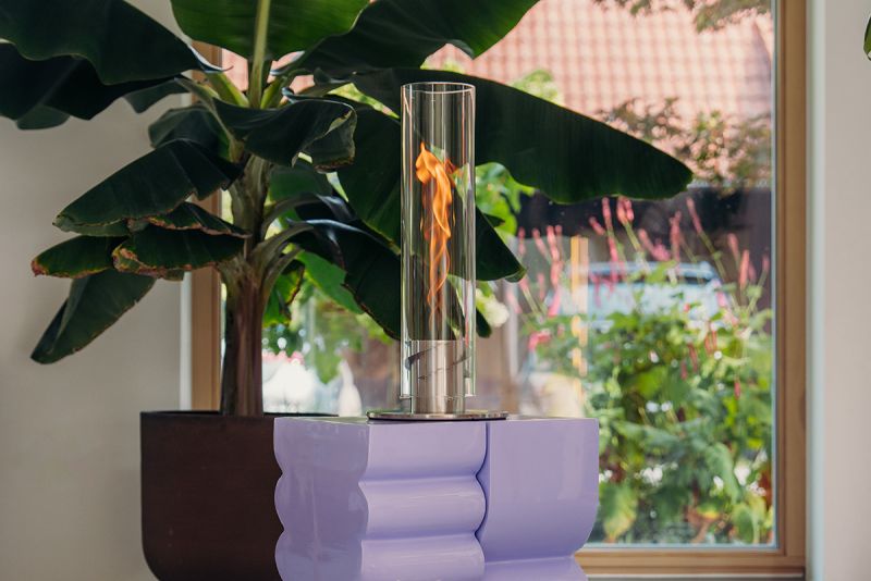 Une cheminée de table Höfats SPIN Classic 900 argent – bioéthanol, au design moderne composé d'un cylindre en verre laissant apparaître la flamme, trône sur un socle ondulé de couleur lavande, au milieu de plantes, près d'une grande fenêtre donnant sur une végétation luxuriante.