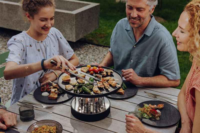 Trois personnes sont assises à une table en plein air, souriantes, en train de faire cuire des légumes et de la viande sur le CIRCLE 35 au bioéthanol avec plancha – Ø 35 cm – Gril de table höfats. Des assiettes remplies de nourriture et des verres sont posés sur la table en bois, créant une ambiance décontractée et détendue dans un jardin verdoyant.