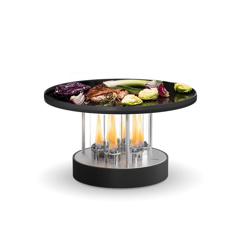 CIRCLE 35 au bioéthanol avec plancha – Ø 35 cm – Gril de table Höfats présenté en situation d'utilisation : un gril de table circulaire sur lequel cuisent divers légumes et viandes sur sa surface noire, avec des flammes visibles qui chauffent la plancha par le dessous, à l'intérieur de son socle cylindrique moderne.