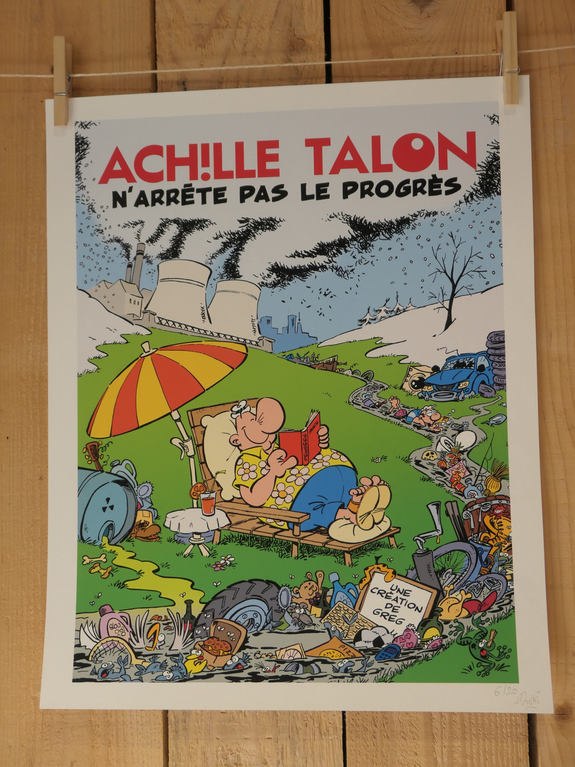 Une lithographie aux couleurs vives intitulée « Lithographie Moski – Achille Talon n’arrête pas le progrès – 40 × 50 cm » représente un personnage de bande dessinée se prélassant tranquillement dans une chaise longue sous un parasol, apparemment indifférent à la pollution qui l’entoure, avec en arrière-plan des usines et des déchets éparpillés. L’œuvre met en avant la phrase : « Achille Talon n’arrête pas le progrès. ».