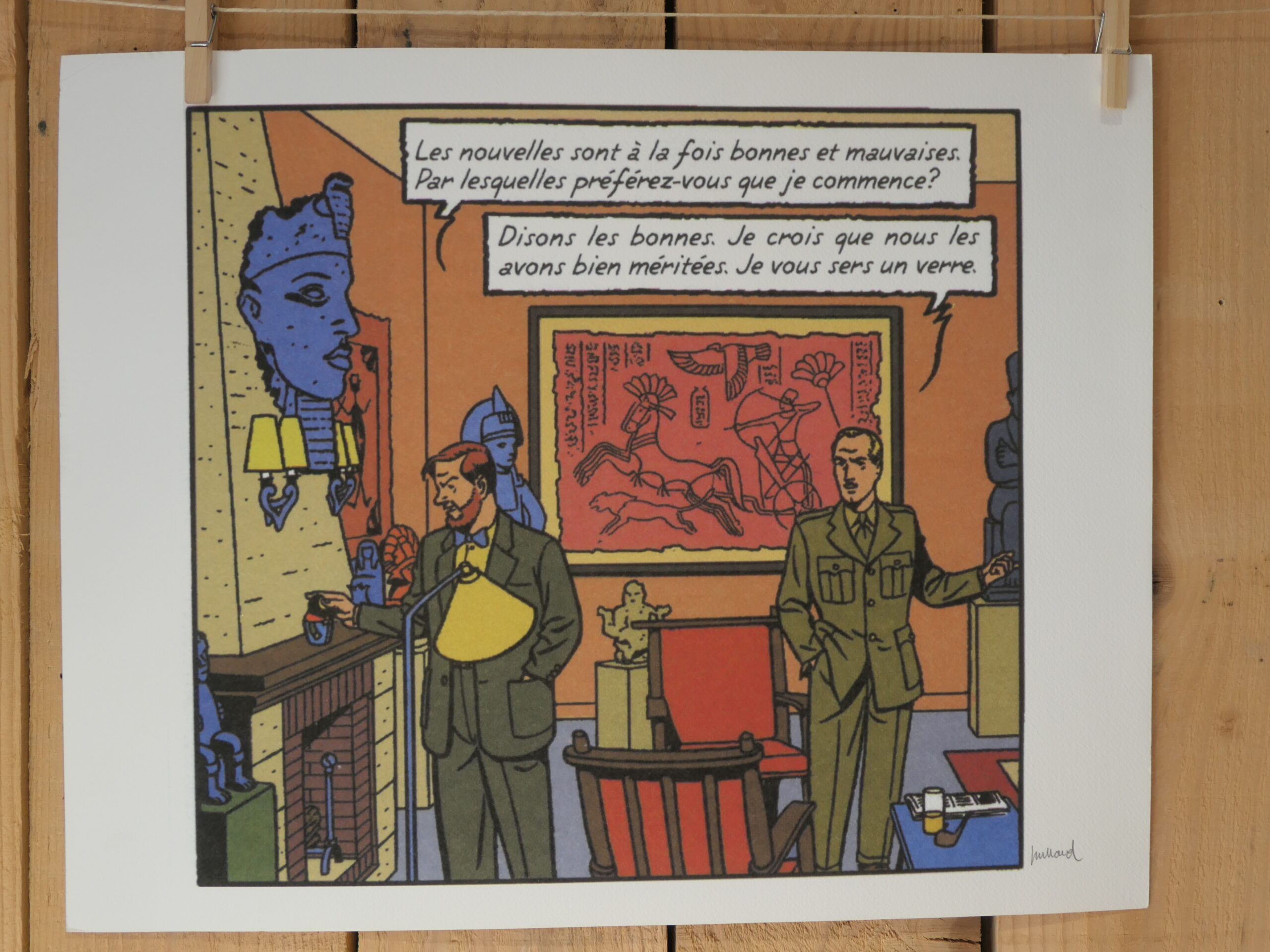 Deux hommes en costume, Blake et Mortimer, discutent dans une pièce ornée d’objets anciens et d’œuvres d’art. L’un d’eux tient un verre à la main, tandis qu’un texte en français apparaît au-dessus d’eux dans une bulle de bande dessinée. L’arrière-plan met en valeur des statues et un imposant relief rouge qui orne le mur. Cette scène figure sur la lithographie « André Juillard – Blake et Mortimer : Les Nouvelles » (40 × 50 cm).