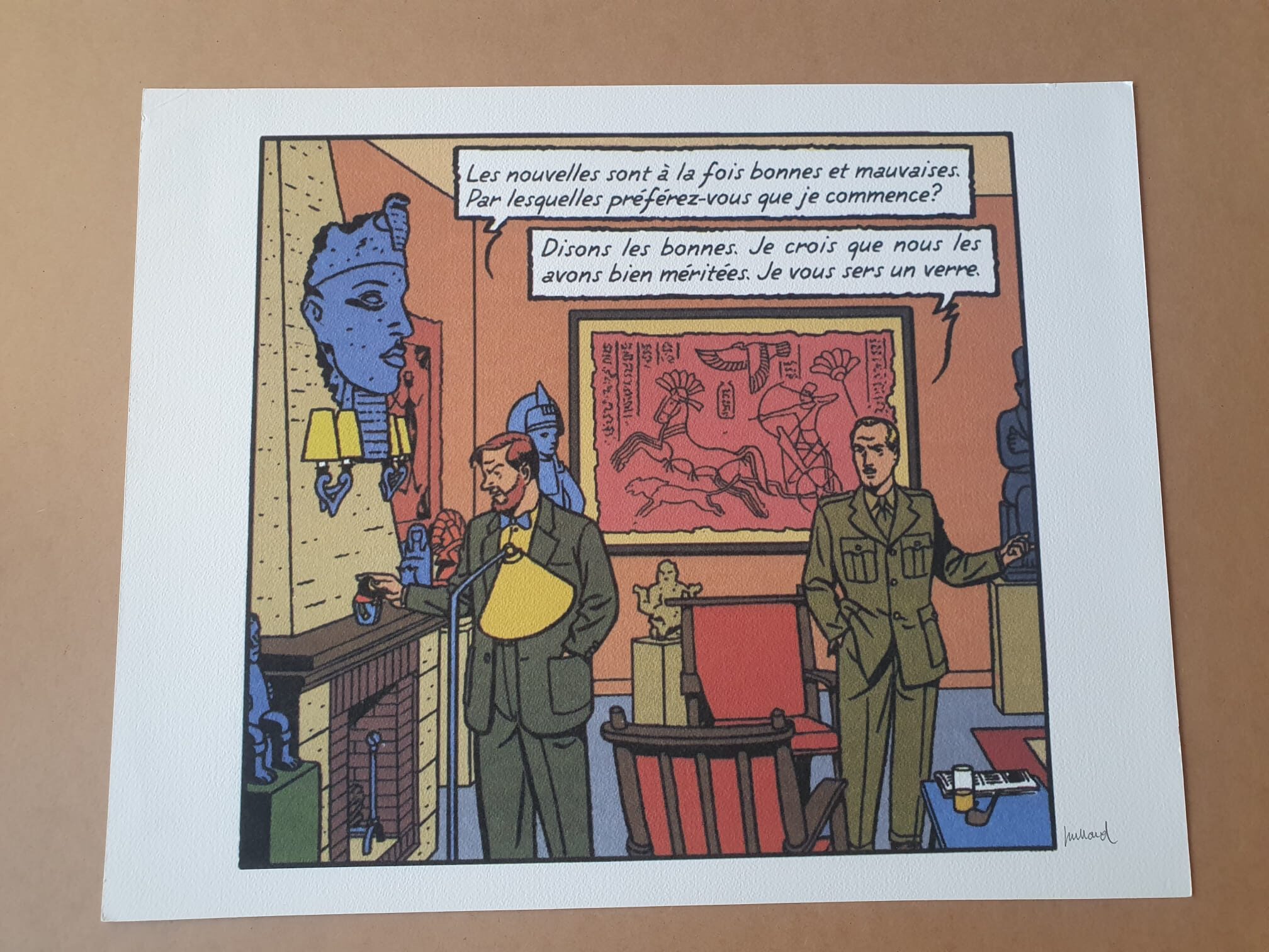 Deux hommes en costume se tiennent dans une pièce décorée d’objets anciens et de peintures murales rouges, tandis qu’une lithographie d’André Juillard – « Blake et Mortimer : Les Nouvelles » (40 × 50 cm) trône bien en évidence sur le mur. L’un des hommes tient un verre à la main tandis que l’autre désigne une statue bleue ; un dialogue en français apparaît au-dessus d’eux.