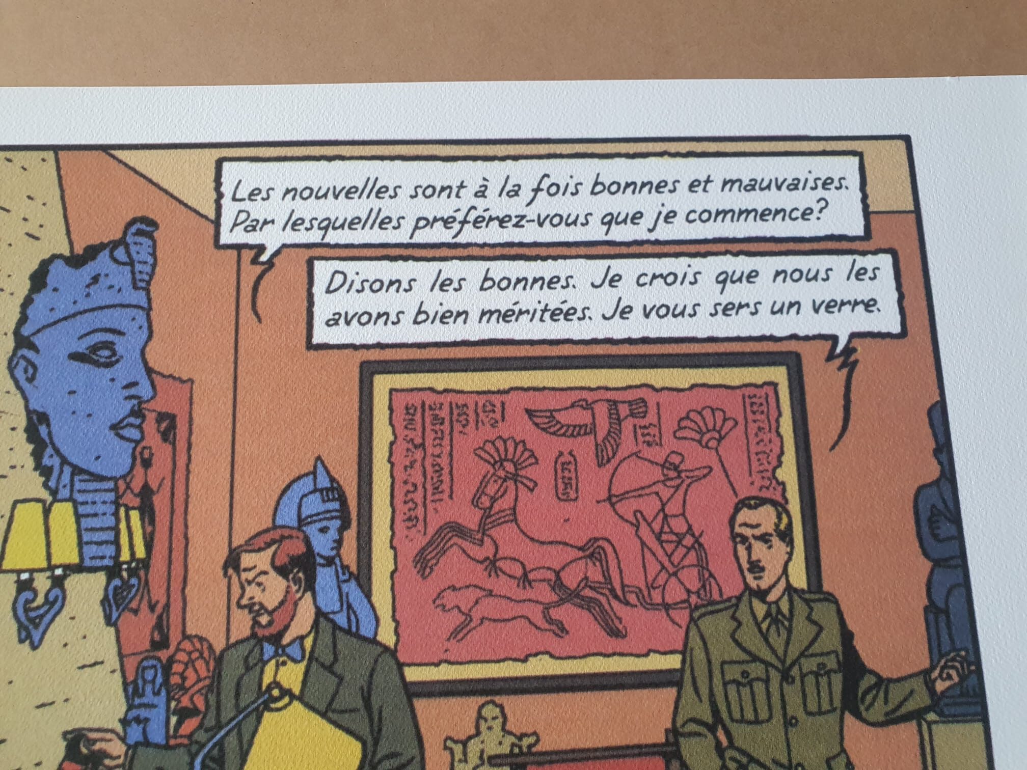 Deux hommes en costume discutent dans une pièce décorée d’œuvres d’art et d’objets de l’Égypte antique, tandis que l’un d’eux tient un dossier. Sur le mur derrière eux est accrochée la « Lithographie André Juillard – Blake et Mortimer : Les Nouvelles – 40 × 50 cm », et leur conversation en français est représentée dans une bulle.