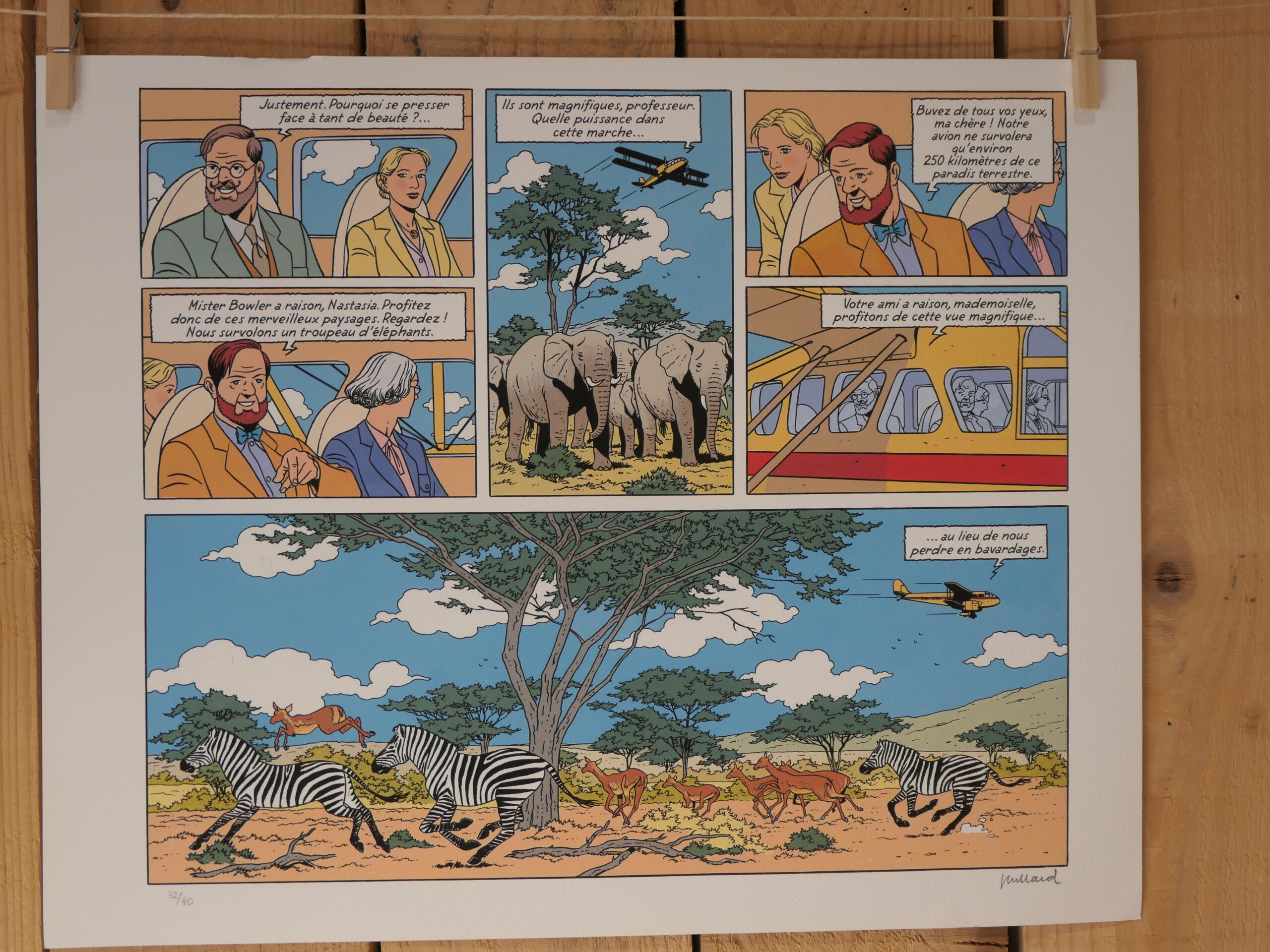 Une lithographie de bande dessinée intitulée « André Juillard – Blake et Mortimer : Planche – 40 × 50 cm » représente, dans ses cases, des éléphants, des zèbres et des antilopes dans la savane africaine, un avion jaune survolant la scène, ainsi que des personnages en train de discuter à l’intérieur de l’appareil.