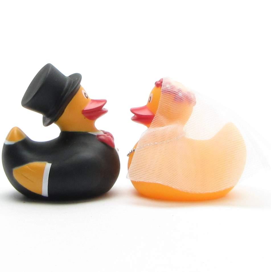 Deux figurines de canards en caoutchouc, habillées en mariés, se font face. Le canard marié porte un smoking noir et un haut-de-forme, tandis que la canarde mariée porte un voile. Tous deux ont un bec orange et sont placés sur un fond blanc.
