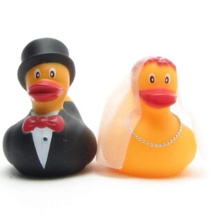 Deux canards en caoutchouc déguisés en mariés : le marié porte un smoking noir et un haut-de-forme, tandis que la mariée porte un voile blanc et un collier de perles.